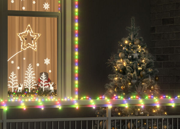 Εξωτερική χριστουγεννιάτικη σκηνή με δέντρο διακοσμημένο με χρυσές μπάλες και φώτα δίπλα σε παράθυρο που περιβάλλεται από πολύχρωμα λαμπάκια. Το παράθυρο έχει φωτεινά στολίδια με αστέρια, νιφάδες χιονιού και σχήματα δέντρων, ενώ στο διπλανό παράθυρο κρέμεται στεφάνι με κόκκινη κορδέλα.